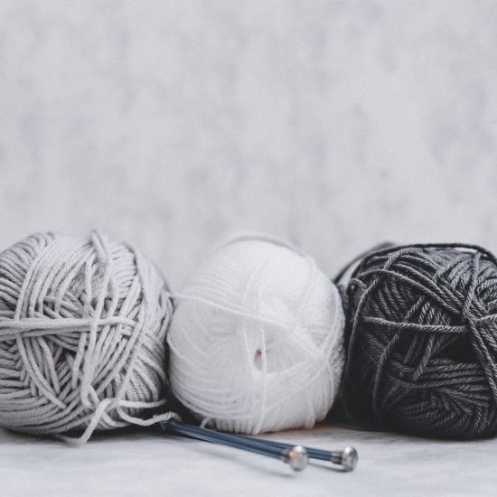 Tres ovillos de lana en color blanco, gris y negro sobre fondo blanco y con dos ajugas de hacer punto. El punch needling es uno de los hobbies y habilidades que quiero probar