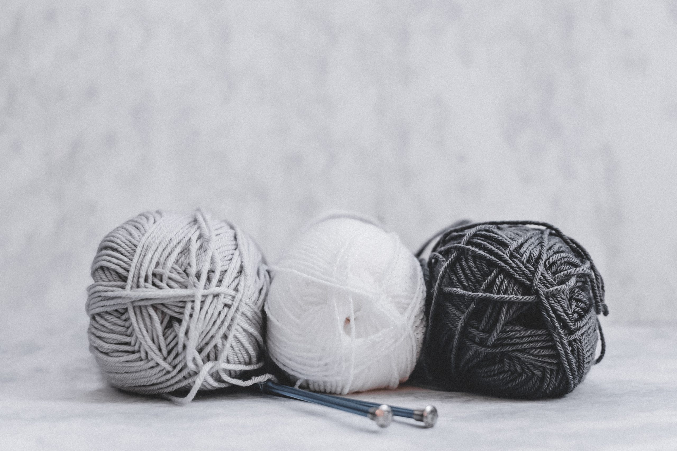 Tres ovillos de lana en color blanco, gris y negro sobre fondo blanco y con dos ajugas de hacer punto. El punch needling es uno de los hobbies y habilidades que quiero probar